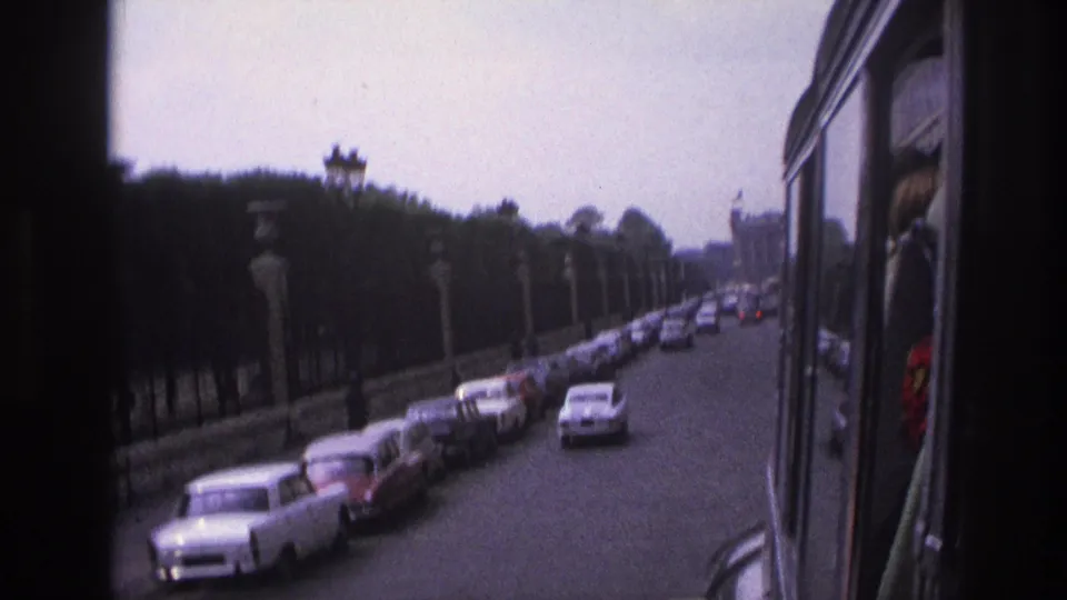 1967 Paris street receding into the distance, with parked cars, moving sedans, a vehicle window edge, trees, stone posts, and iron fencing.