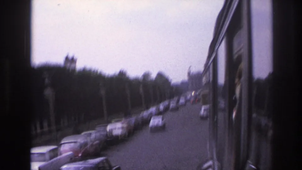 1967 Paris street traffic viewed from a moving vehicle, with parked cars, trees, and dark window framing.