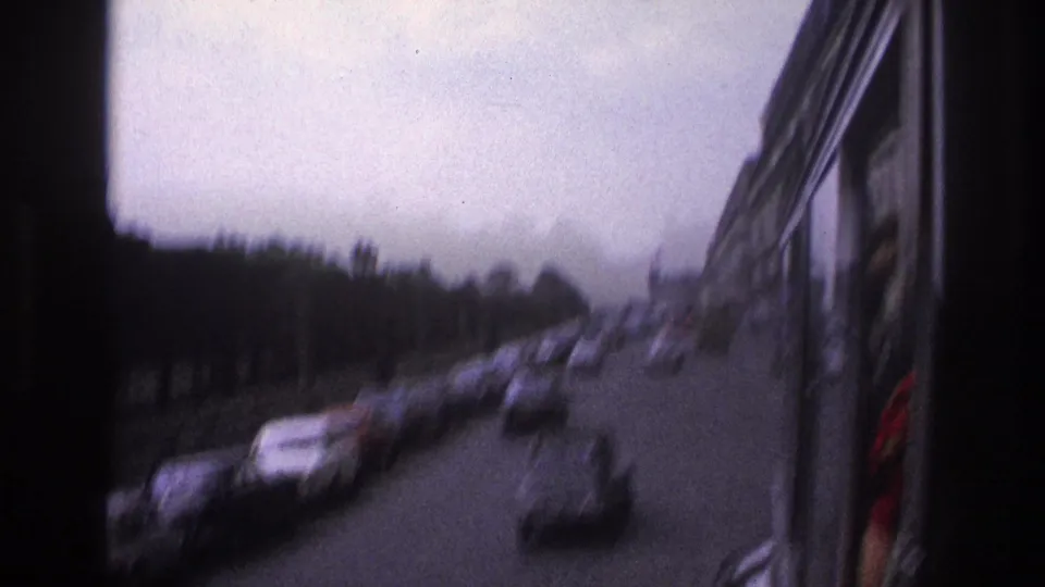 Soft 8mm view of 1967 Paris street traffic, with cars, trees, and the side of the filming vehicle visible.