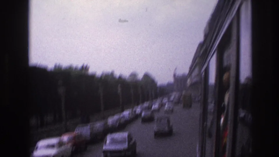 1967 Paris street view from a moving vehicle, showing traffic lanes, parked cars, trees, and muted overcast light.