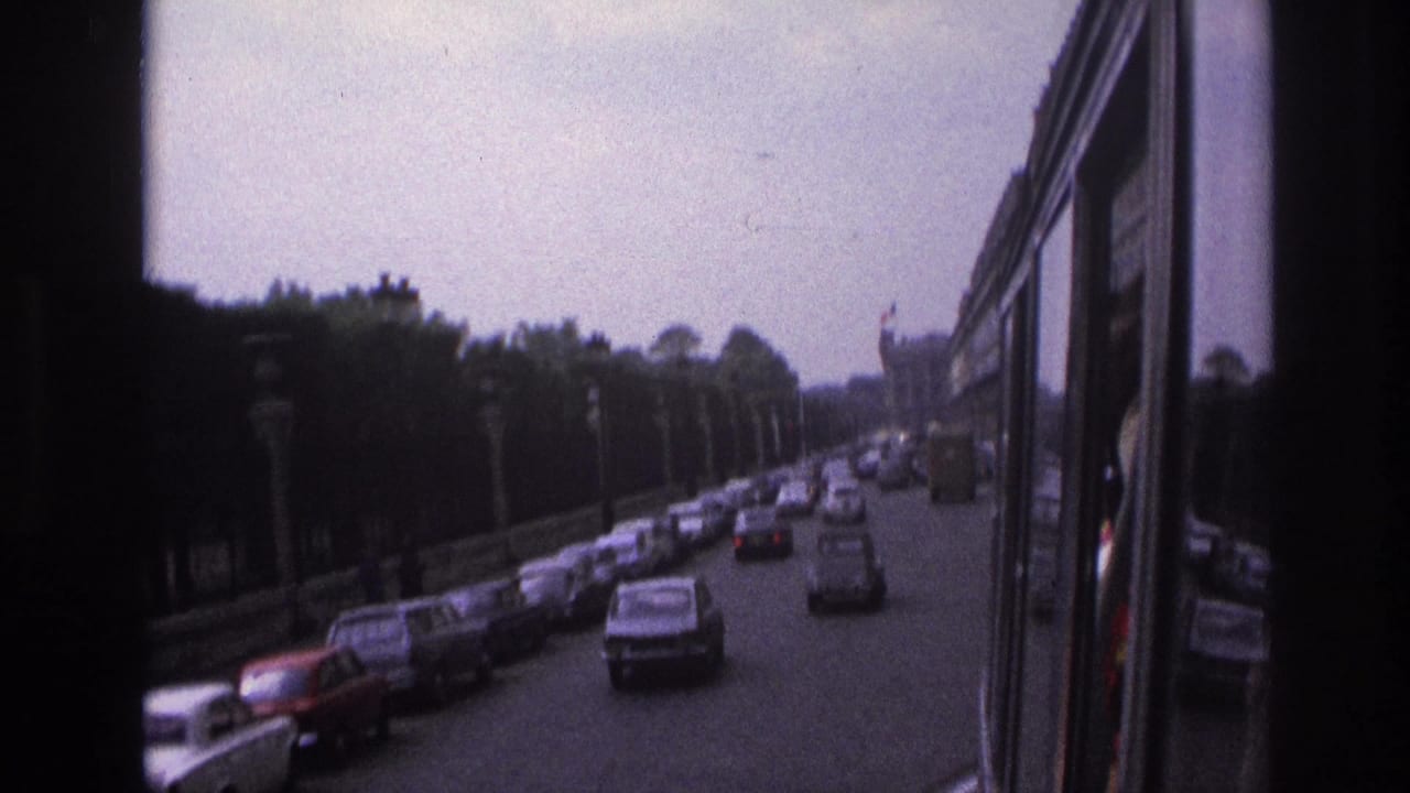 1967 Paris boulevard traffic viewed from a moving bus, with classic cars, lamps, trees, and a distant formal building.
