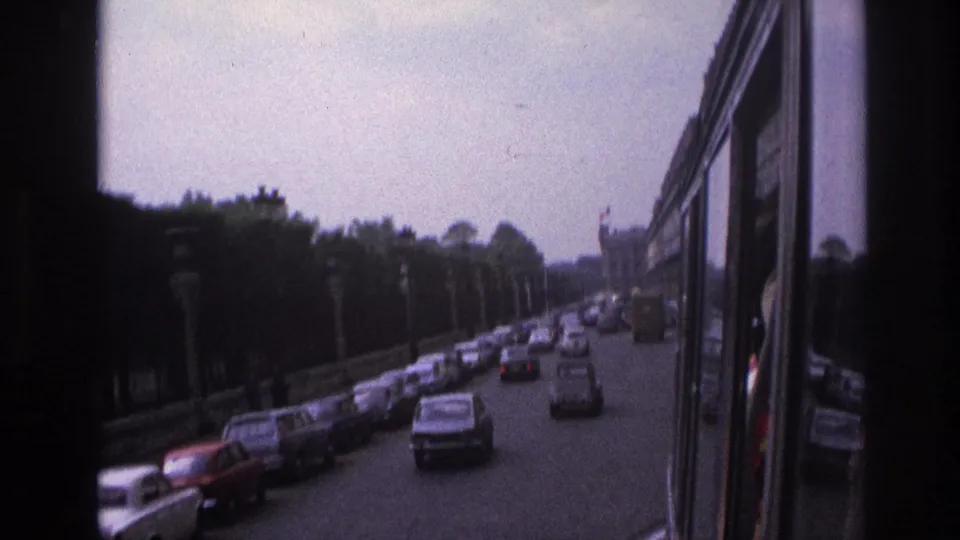 1967 Paris boulevard traffic viewed from a moving bus, with classic cars, lamps, trees, and a distant formal building.