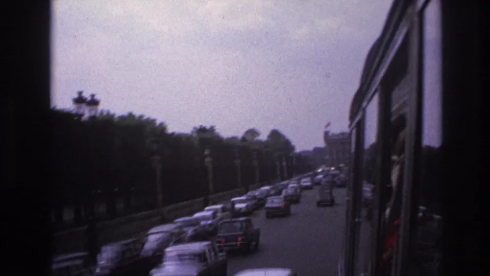 Dark 8mm frame of a Paris boulevard from a bus, showing classic cars, trees, lamps, and muted evening light.