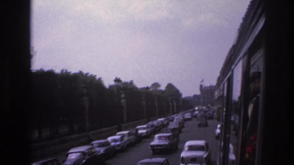 1967 Paris bus-side view with a stream of classic cars, roadside trees, lamps, and a distant building shape.