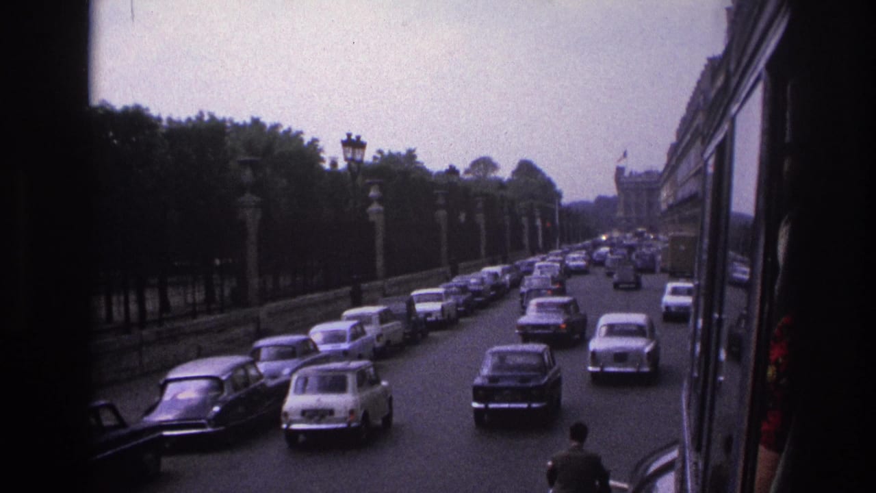 1967 Paris traffic on a broad boulevard, with mid-century cars, trees, stone posts, and a moving-vehicle viewpoint.