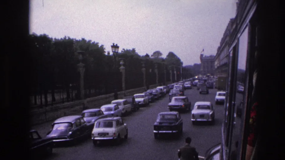1967 Paris traffic on a broad boulevard, with mid-century cars, trees, stone posts, and a moving-vehicle viewpoint.