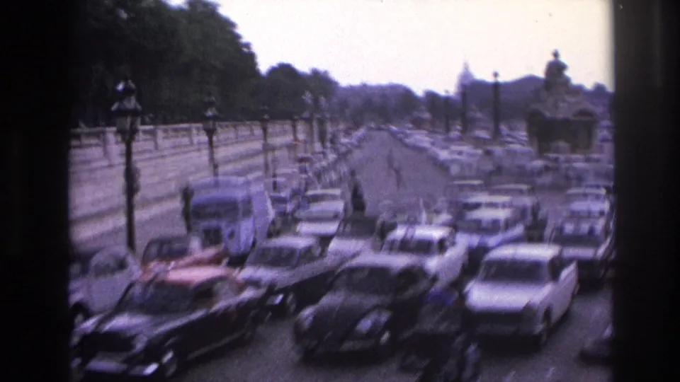 1967 Paris roadway crowded with cars beside stone walls, street lamps, trees, and a blurred moving-camera foreground.