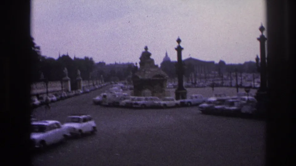 1967 Paris traffic circles near a sculptural monument, with parked and moving cars, lamps, trees, and a distant dome.