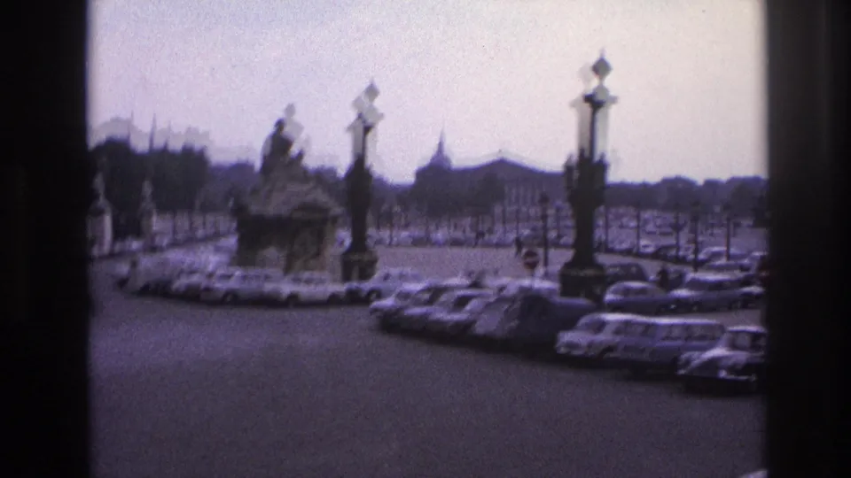 1967 Paris home-movie frame showing an ornate fountain, decorative lampposts, and parked cars viewed through a window frame.