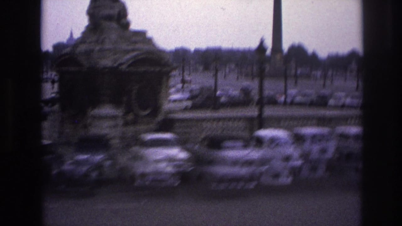 Blurred 1967 Paris frame with a fountain structure, parked cars, and formal lamp posts seen through a window.