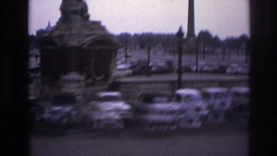 Blurred 1967 Paris frame with a fountain structure, parked cars, and formal lamp posts seen through a window.