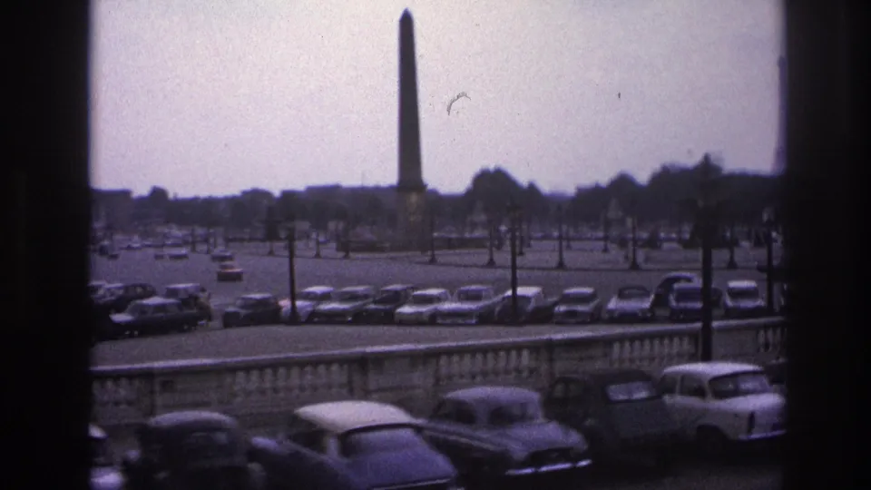 1967 Paris view of an obelisk across a broad parking area, with a stone balustrade and classic cars in the foreground.