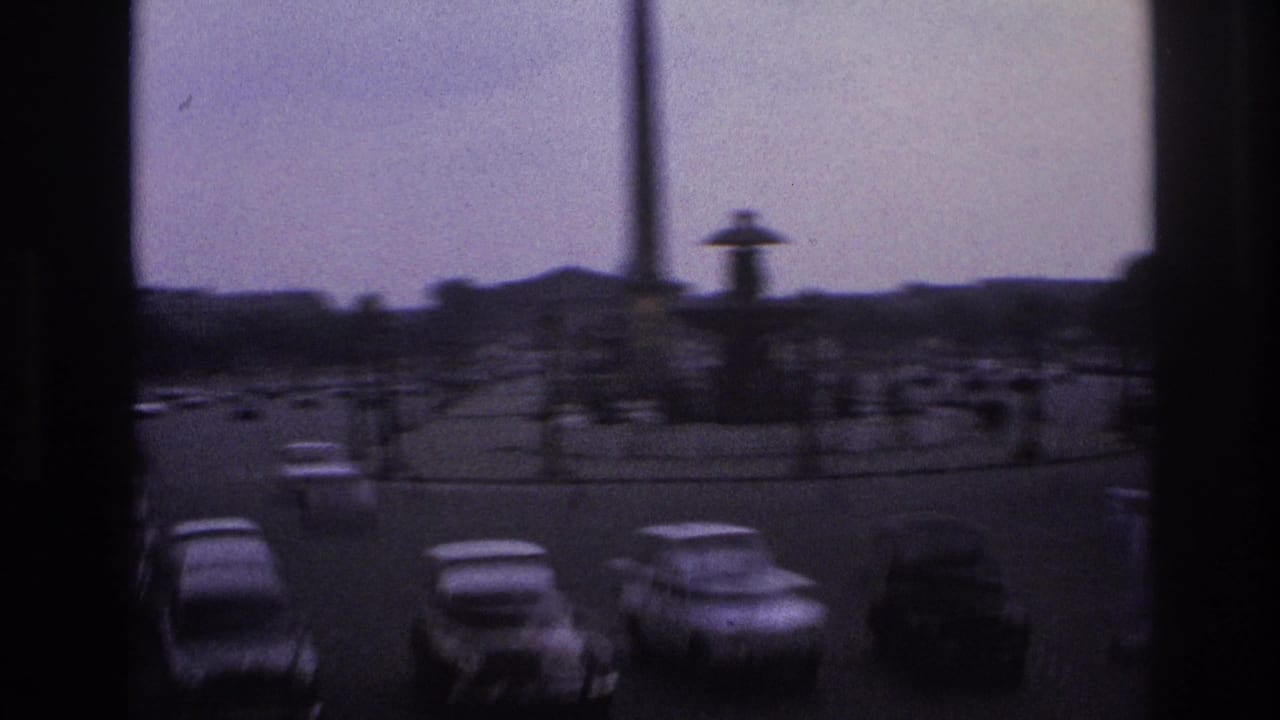 1967 Paris street view from behind window frames, with cars circling a monument under an overcast sky.