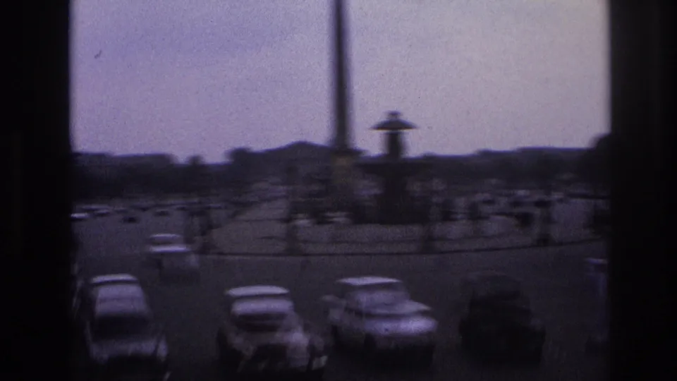 1967 Paris street view from behind window frames, with cars circling a monument under an overcast sky.