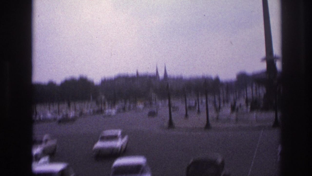 Cars travel through a broad roundabout with bare trees, lampposts, and spires in the distance.