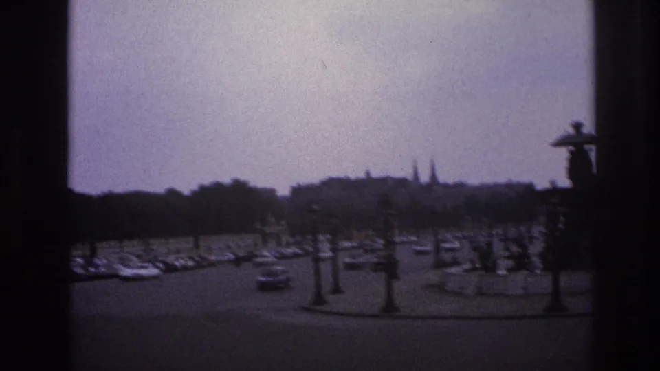 Visitor-shot 1967 Paris view of a traffic circle with moving cars, bare trees, and distant rooftops softened by 8mm grain.