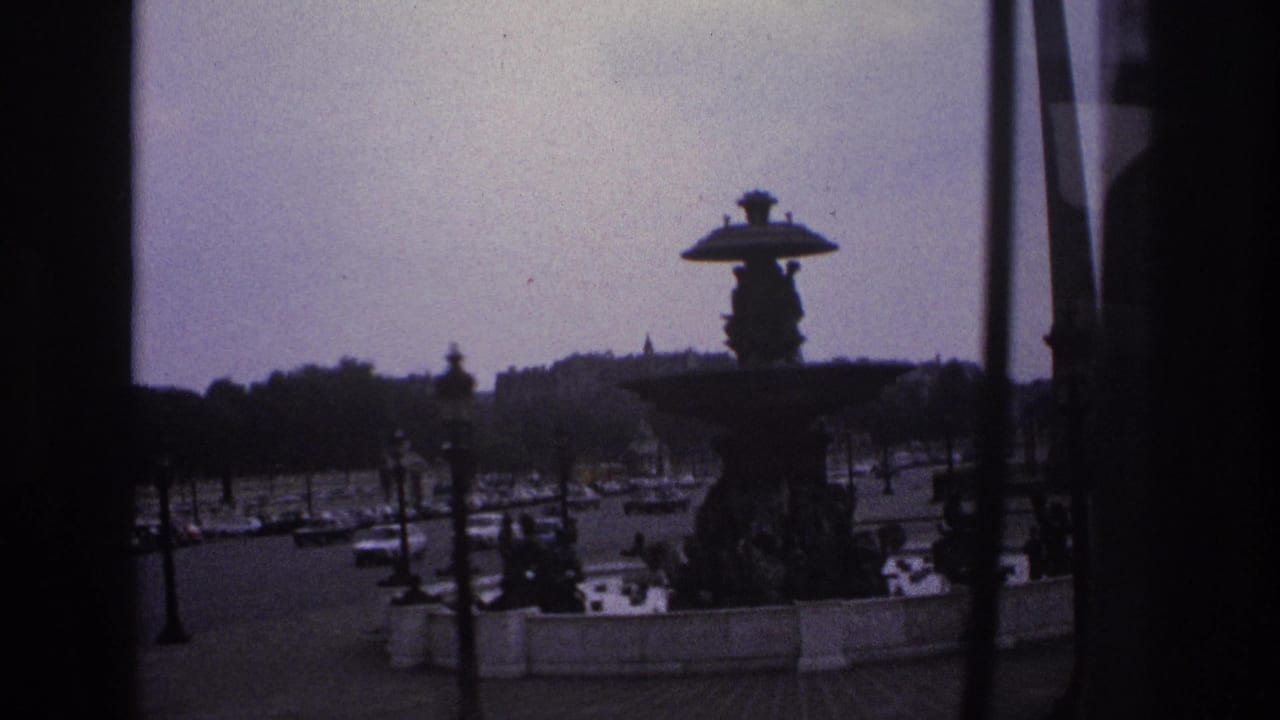 1967 Paris home-movie frame showing an ornate fountain, traffic, pedestrians, lamps, and distant trees through a dark window frame.