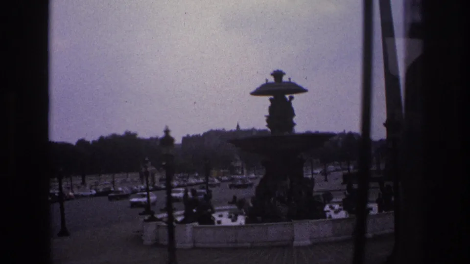 1967 Paris home-movie frame showing an ornate fountain, traffic, pedestrians, lamps, and distant trees through a dark window frame.
