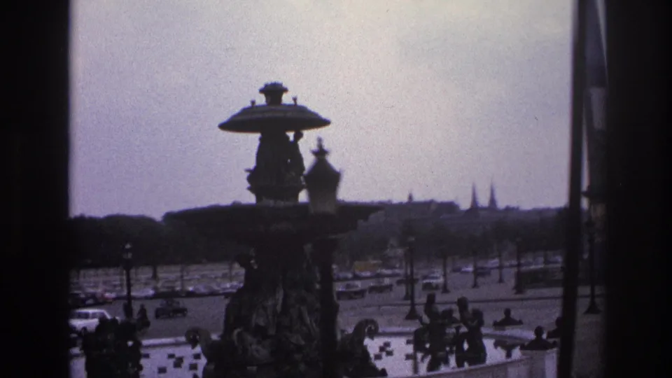 1967 Paris street scene with the fountain silhouetted against a bright sky, cars moving through the square, and people near the plaza edge.