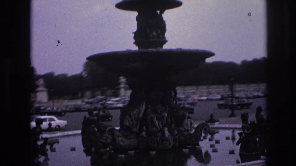 Close 1967 Paris home-movie view of the fountain basin and sculptural figures, with cars, benches, pedestrians, and lamps in the square.