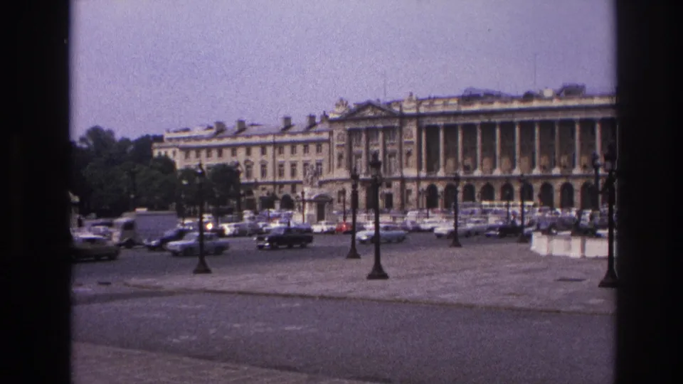 1967 Paris frame showing a grand stone building with columns, parked and moving cars, formal lamps, and a broad paved public square.