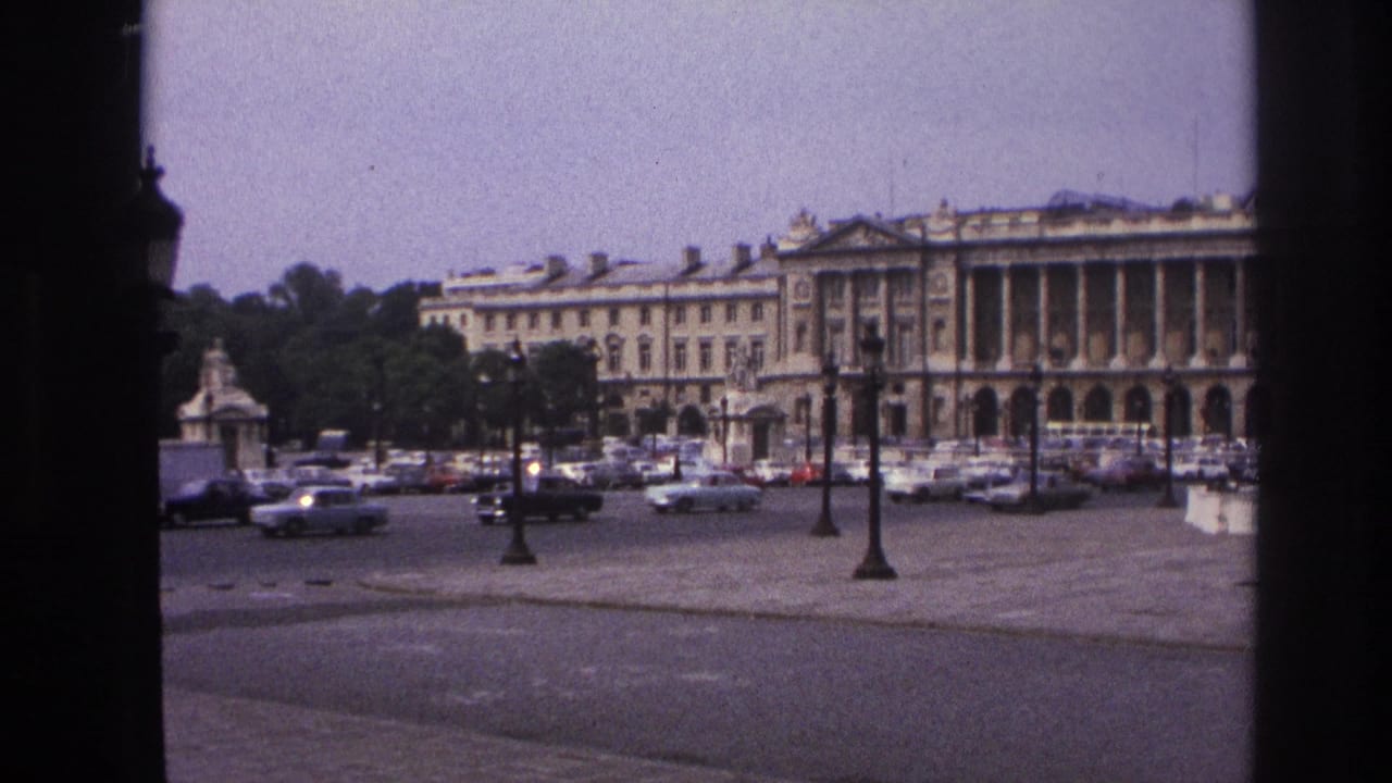 1967 Paris home-movie frame showing a grand colonnaded building across a paved square with classic cars and ornate lampposts.