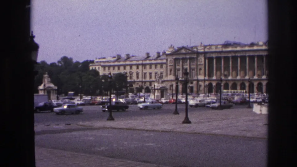 1967 Paris home-movie frame showing a grand colonnaded building across a paved square with classic cars and ornate lampposts.