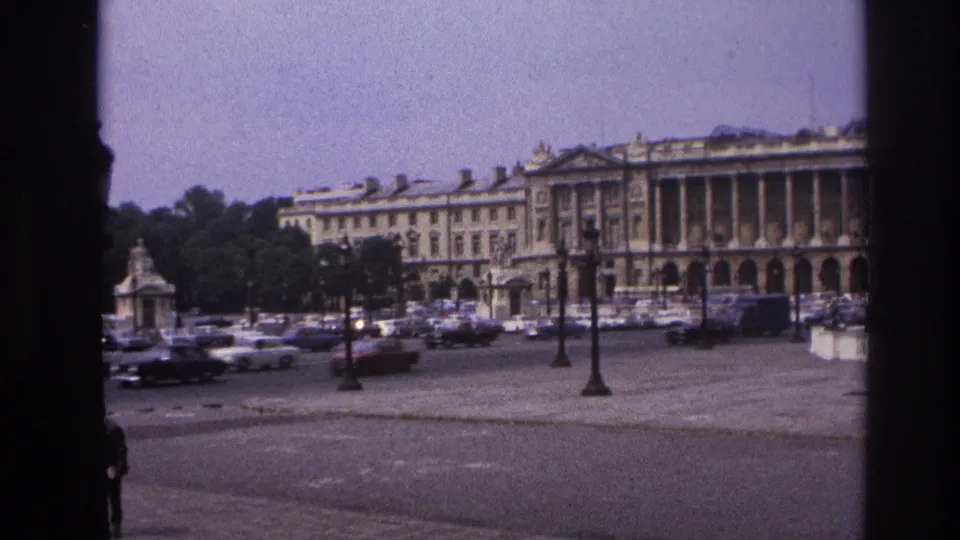 1967 Paris frame with muted color and grain, showing traffic and parked cars before a grand classical building.