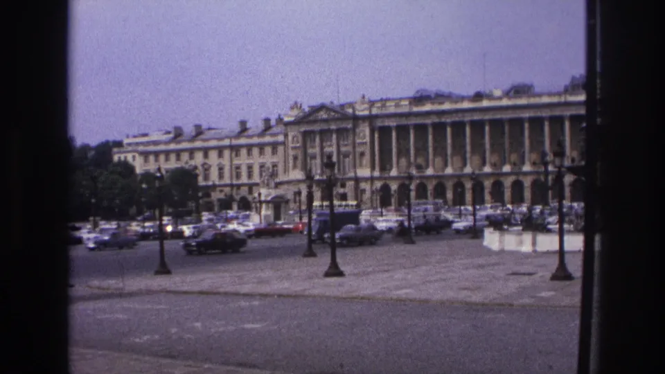 1967 Paris home-movie still of classic cars moving across a broad plaza before monumental stone architecture.