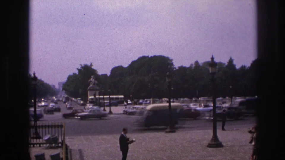 1967 Paris home-movie frame looking across a busy square with classic cars, pedestrians, trees, lamp posts, and a monument.