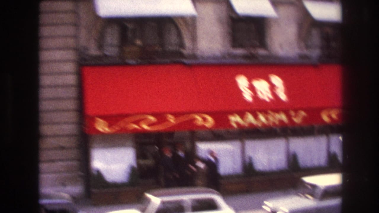 1967 Paris street view with a red Maxim's awning, stone facade, pedestrians, and passing cars, softened by motion blur.