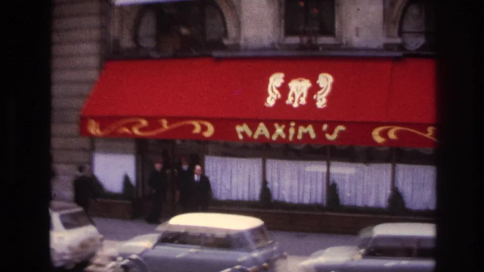 1967 Paris frame showing the red Maxim's restaurant awning, people near the entrance, parked cars, and a stone building exterior.