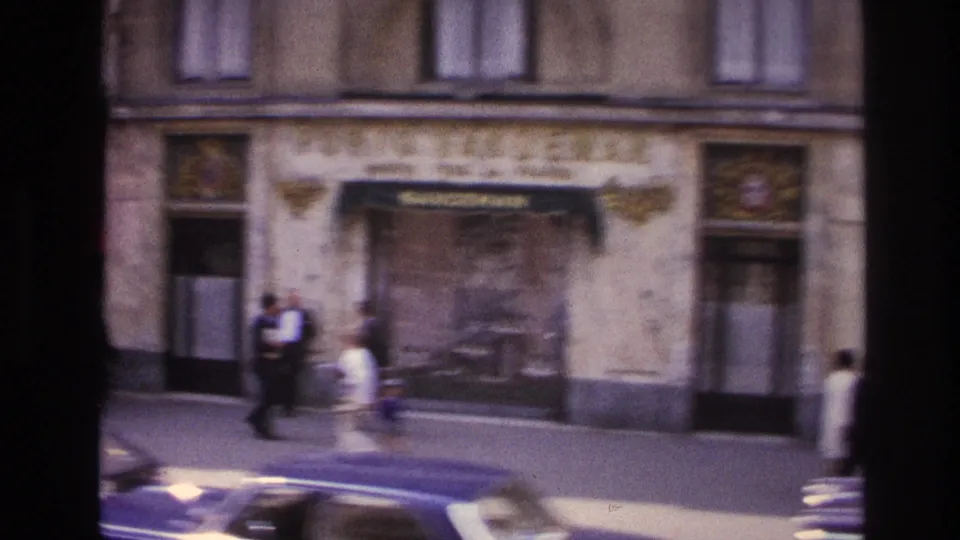 1967 Paris street frame with pedestrians, parked cars, stone storefront details, windows, and a shuttered central entrance.