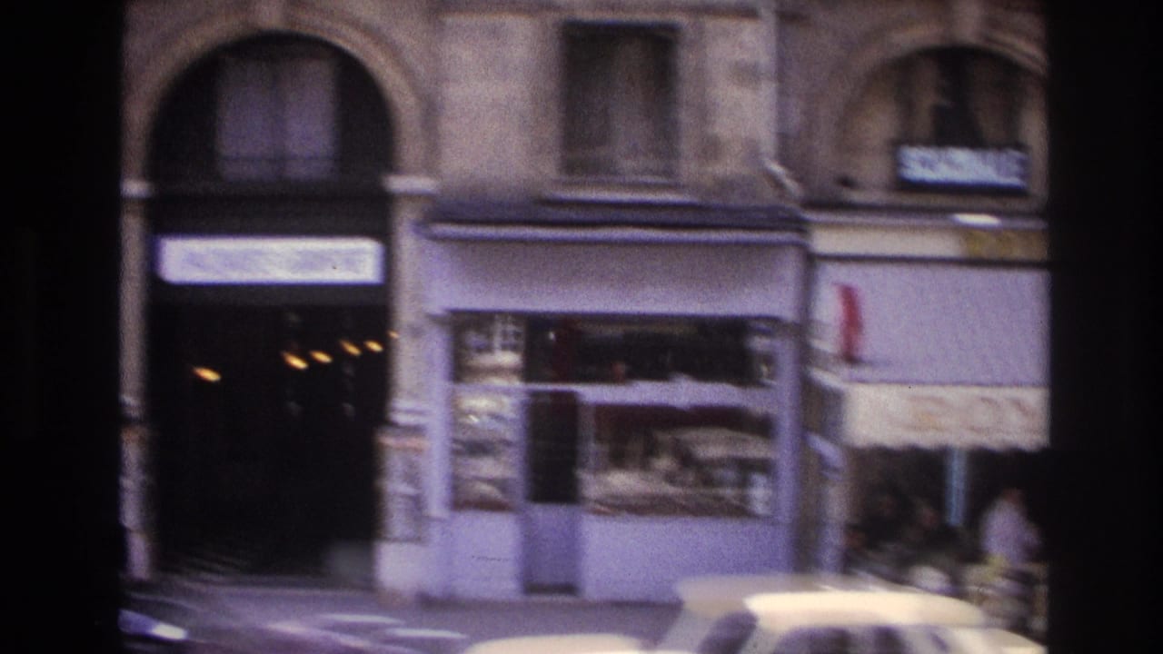 1967 Paris street view with a blurred building facade, shopfronts, and a light-colored car passing in the foreground.