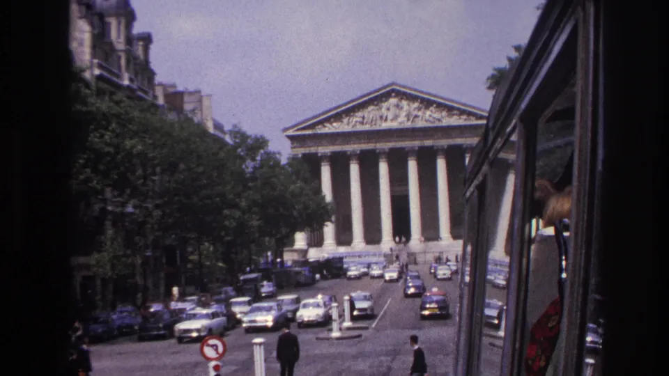 1967 Paris view toward the Panthéon from a moving vehicle, with traffic, pedestrians, trees, and the vehicle edge at right.