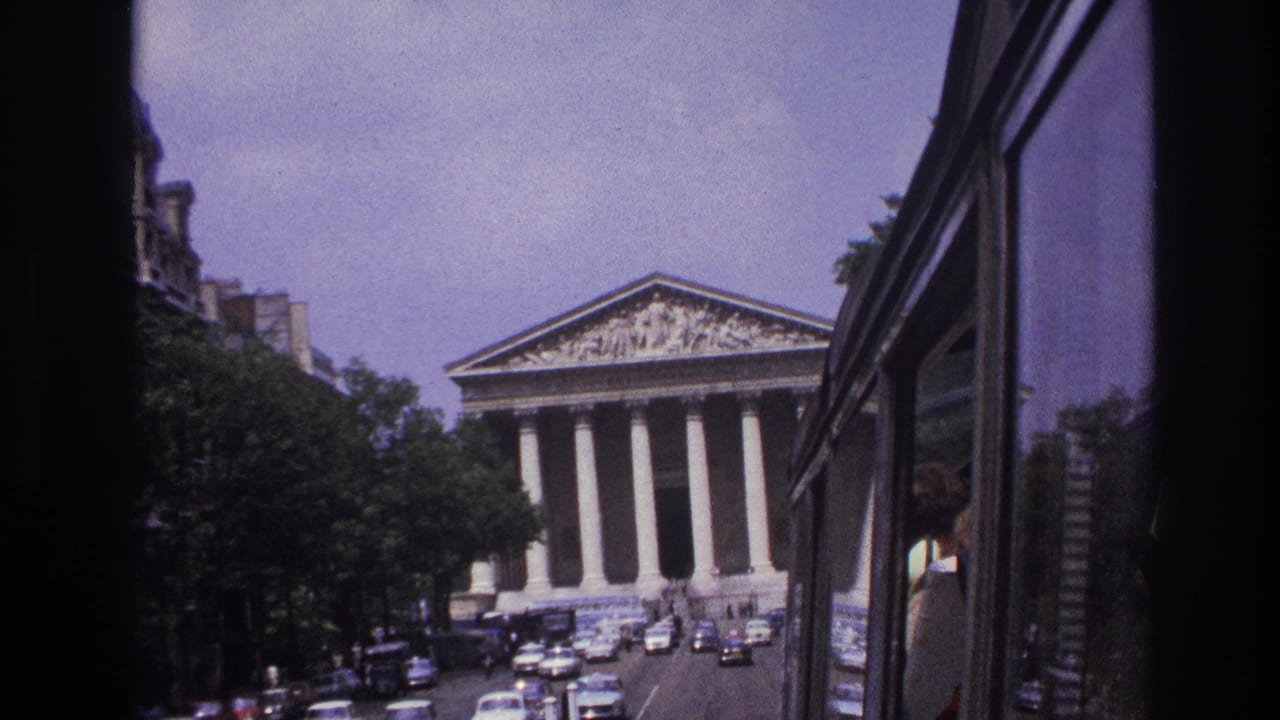 1967 Paris street view from a moving vehicle, with a columned neoclassical landmark ahead and classic cars in traffic.