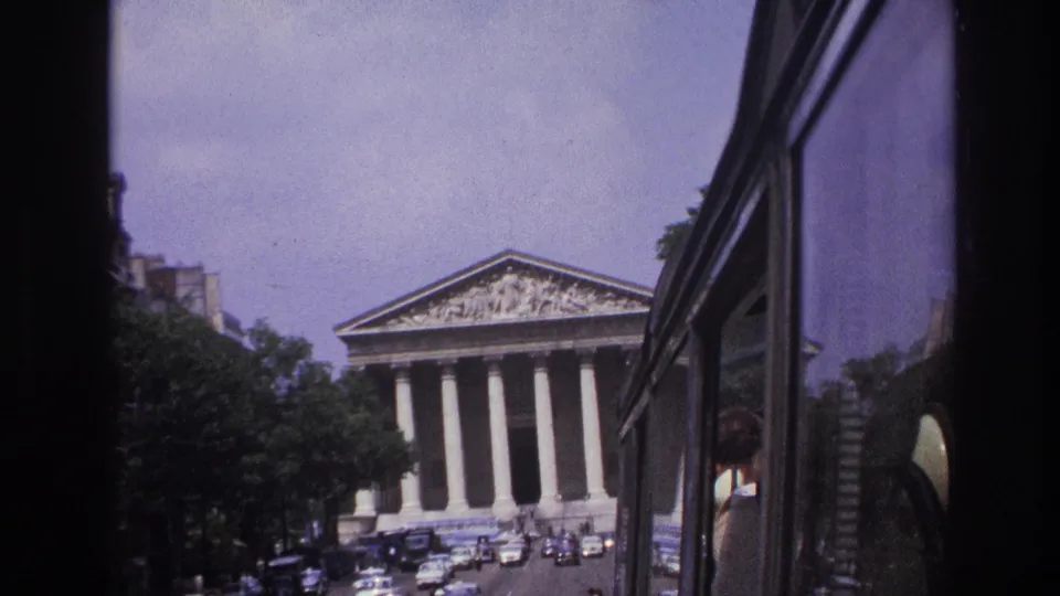 Paris landmark facade with tall columns, seen through a vehicle window beside 1960s cars and street trees.