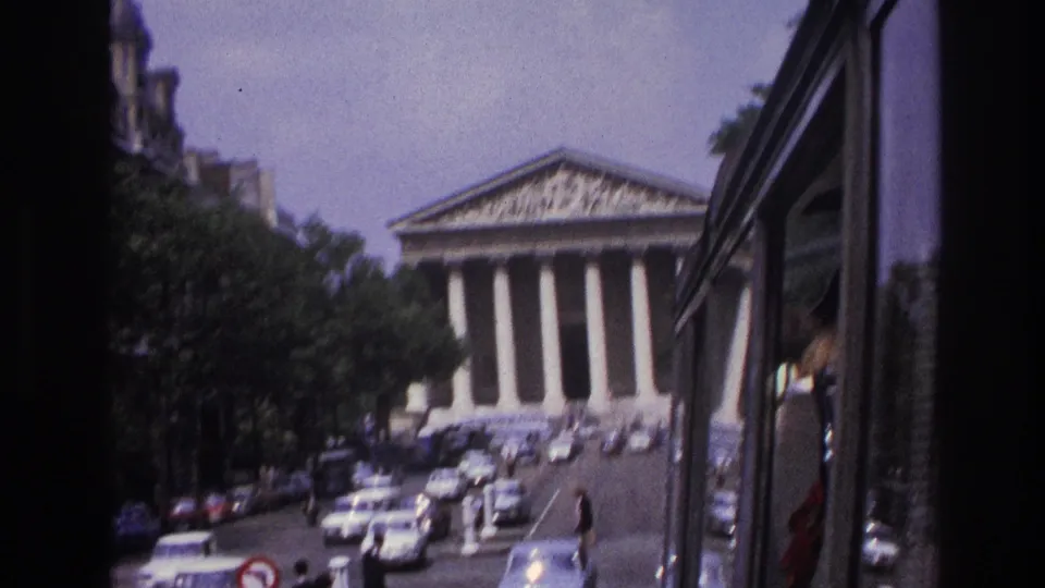 Motion-blurred 1967 Paris street view with a columned landmark, cars, pedestrians, and the vehicle edge in frame.