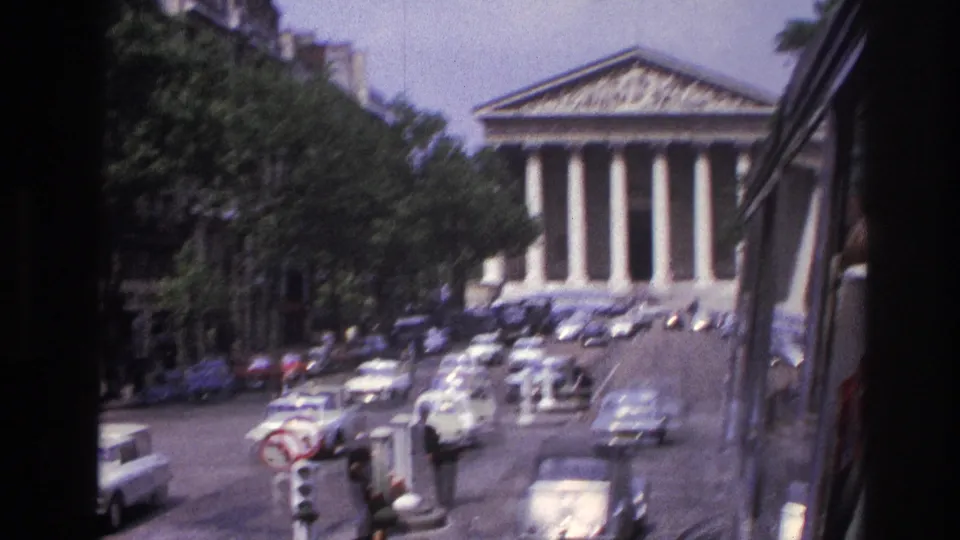 1967 Paris traffic scene with classic cars, a traffic signal, pedestrians, and a neoclassical landmark beyond.