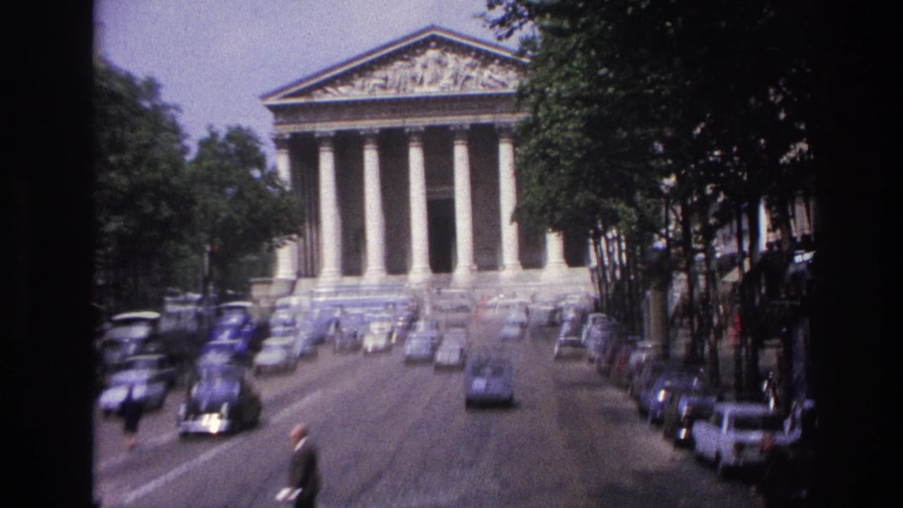 1967 Paris home-movie view of a grand columned civic building seen along a street lined with period cars and trees.