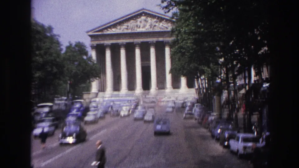 1967 Paris home-movie view of a grand columned civic building seen along a street lined with period cars and trees.