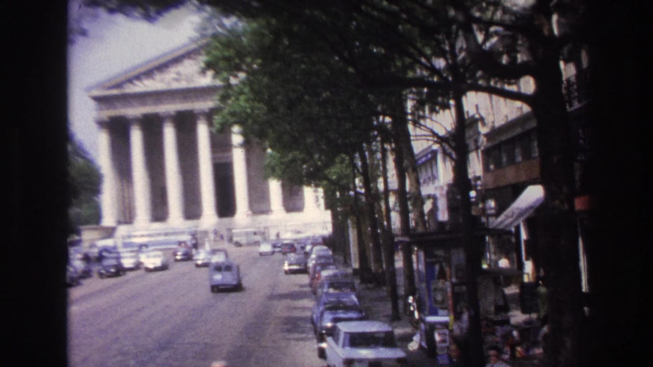 Moving 8mm view toward a Paris columned landmark, with parked cars, trees, and shopfronts along the right side.