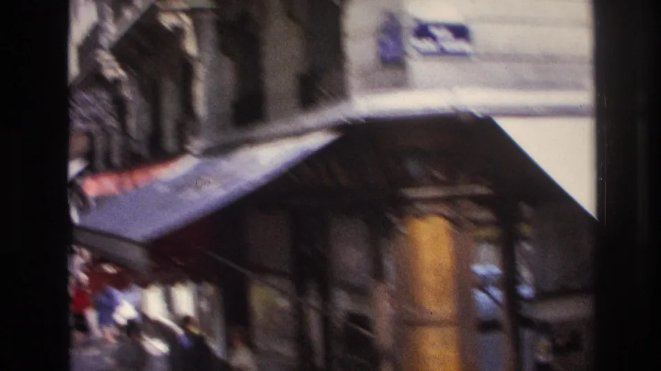 Soft, motion-blurred 1967 Paris street view with an awning, shopfront edge, pedestrians, and passing traffic.