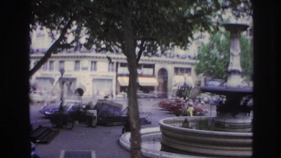 Paris home-movie view of a fountain square with trees, benches, parked cars, pedestrians, and surrounding street architecture.