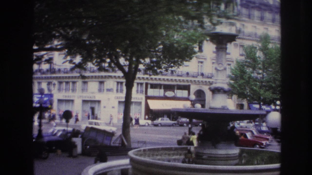 1967 Paris street view across a fountain toward formal buildings, awnings, pedestrians, and period traffic.