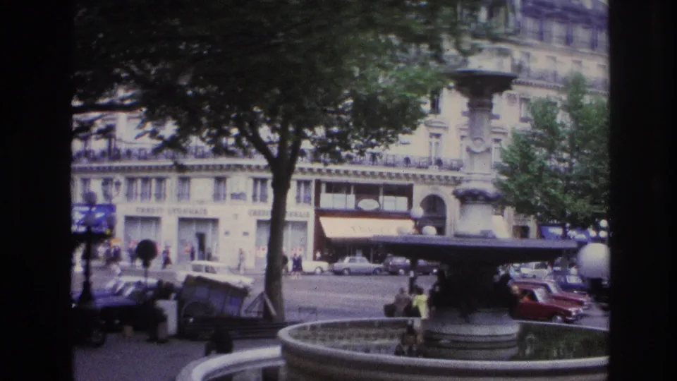 1967 Paris street view across a fountain toward formal buildings, awnings, pedestrians, and period traffic.