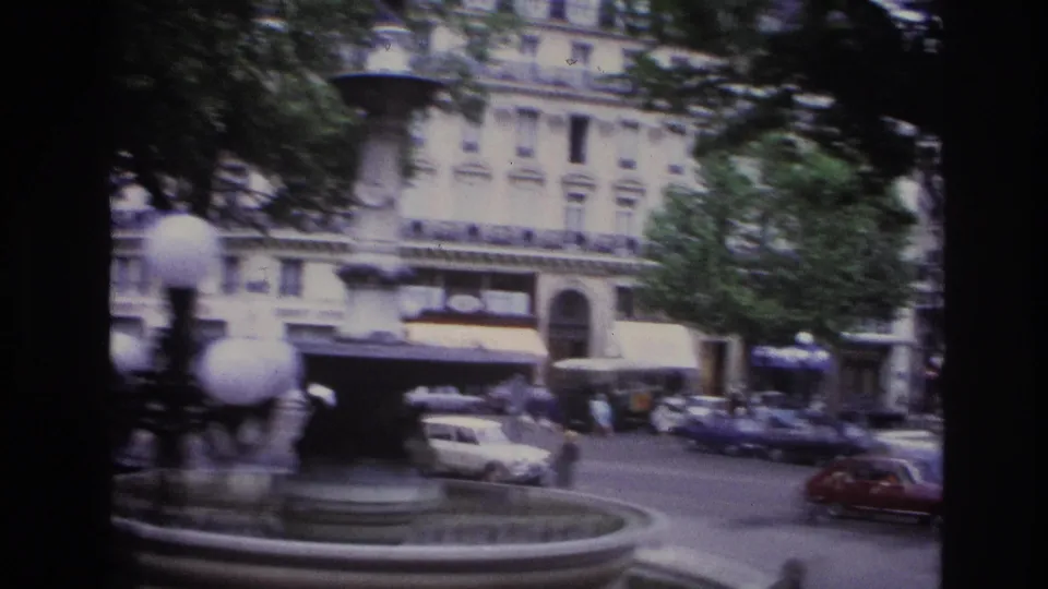 1967 Paris fountain view with soft 8mm motion blur, globe lamps, moving cars, trees, and a building facade.