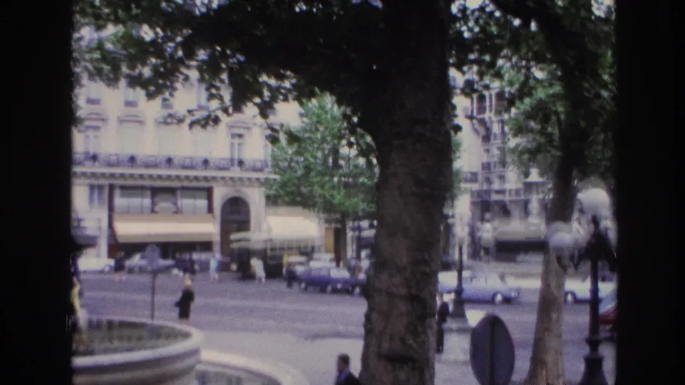 1967 Paris street corner framed by a tree trunk, with a fountain edge, pedestrians, period cars, and awnings across the road.