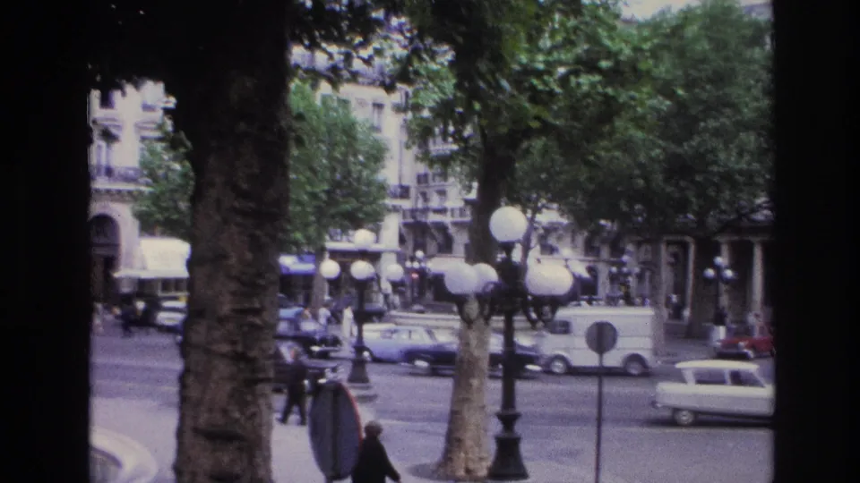 1967 Paris street view with globe lamps by the fountain, leafy trees, pedestrians, period cars, and multi-story buildings.