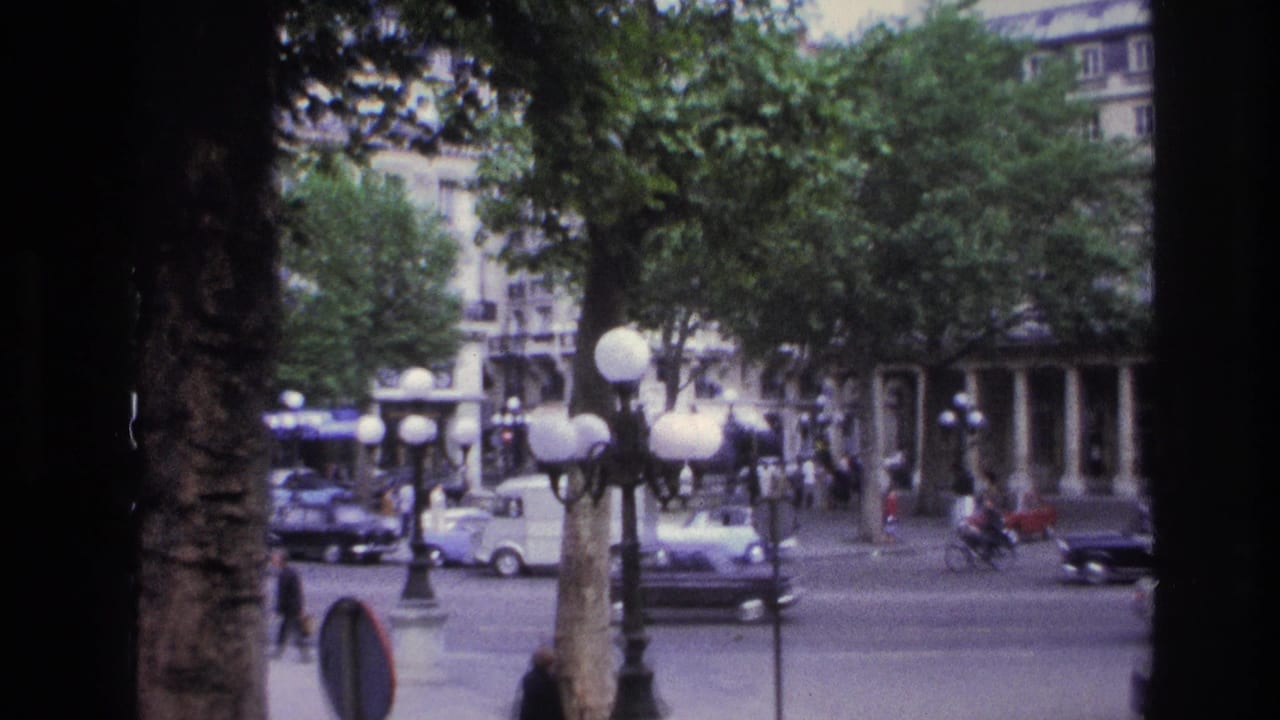 1967 Paris street view through a window frame, showing leafy trees, globe lampposts, classic cars, pedestrians, and multi-story facades.
